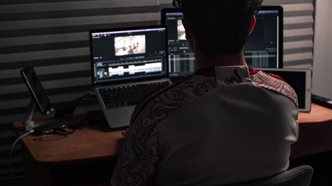 Man sitting at desk working on video editing software with dual monitors in an office.